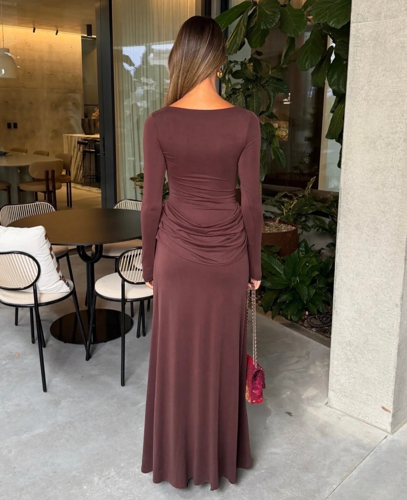 A woman stands with her back to the camera wearing a long-sleeve, floor-length mauve-brown dress with ruching at the waist. She is holding a small pink handbag in an outdoor patio setting with tables, chairs, and green plants.