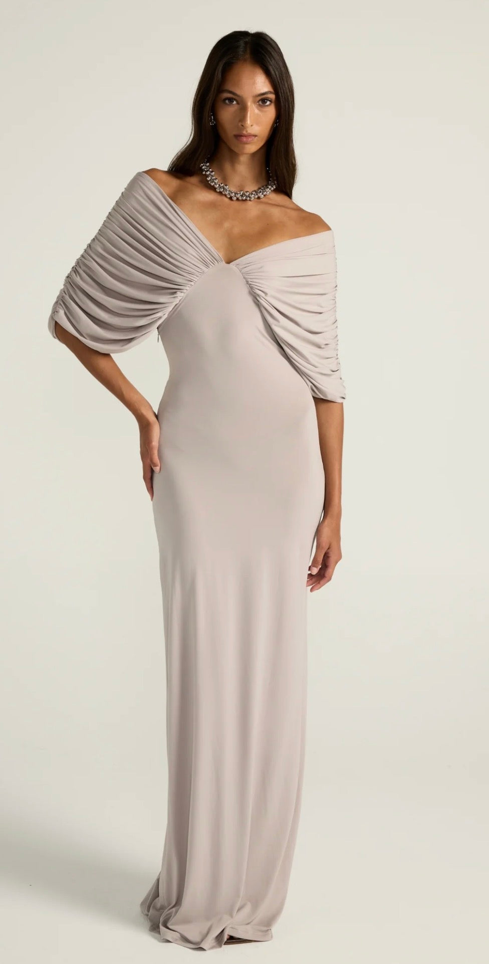 A woman modeling a floor-length off-the-shoulder gown in a light warm gray or greige tone, with draped ruching across the bodice and sleeves, photographed against a plain light background.