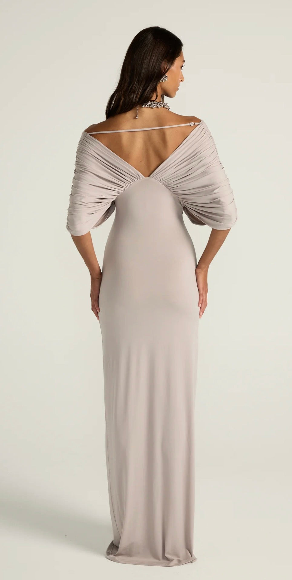A woman modeling a floor-length off-the-shoulder evening gown from the back, featuring draped short sleeves and a low open back, in a light blush-beige color against a plain light gray studio background.