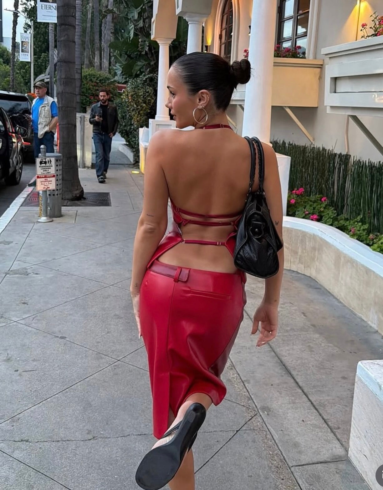 A woman walking on a city sidewalk wearing a bright red backless wrap-style dress with a structured waist and black platform heels, carrying a black shoulder bag.