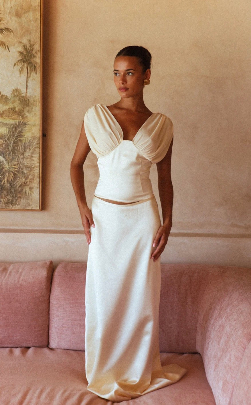 A woman models a cream satin two-piece set consisting of a cropped top with draped puff sleeves and a matching high-waisted skirt, standing on a dusty pink sofa against a textured beige wall.