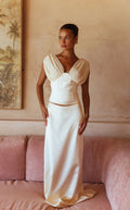 A woman models a cream satin two-piece set consisting of a cropped top with draped puff sleeves and a matching high-waisted skirt, standing on a dusty pink sofa against a textured beige wall.