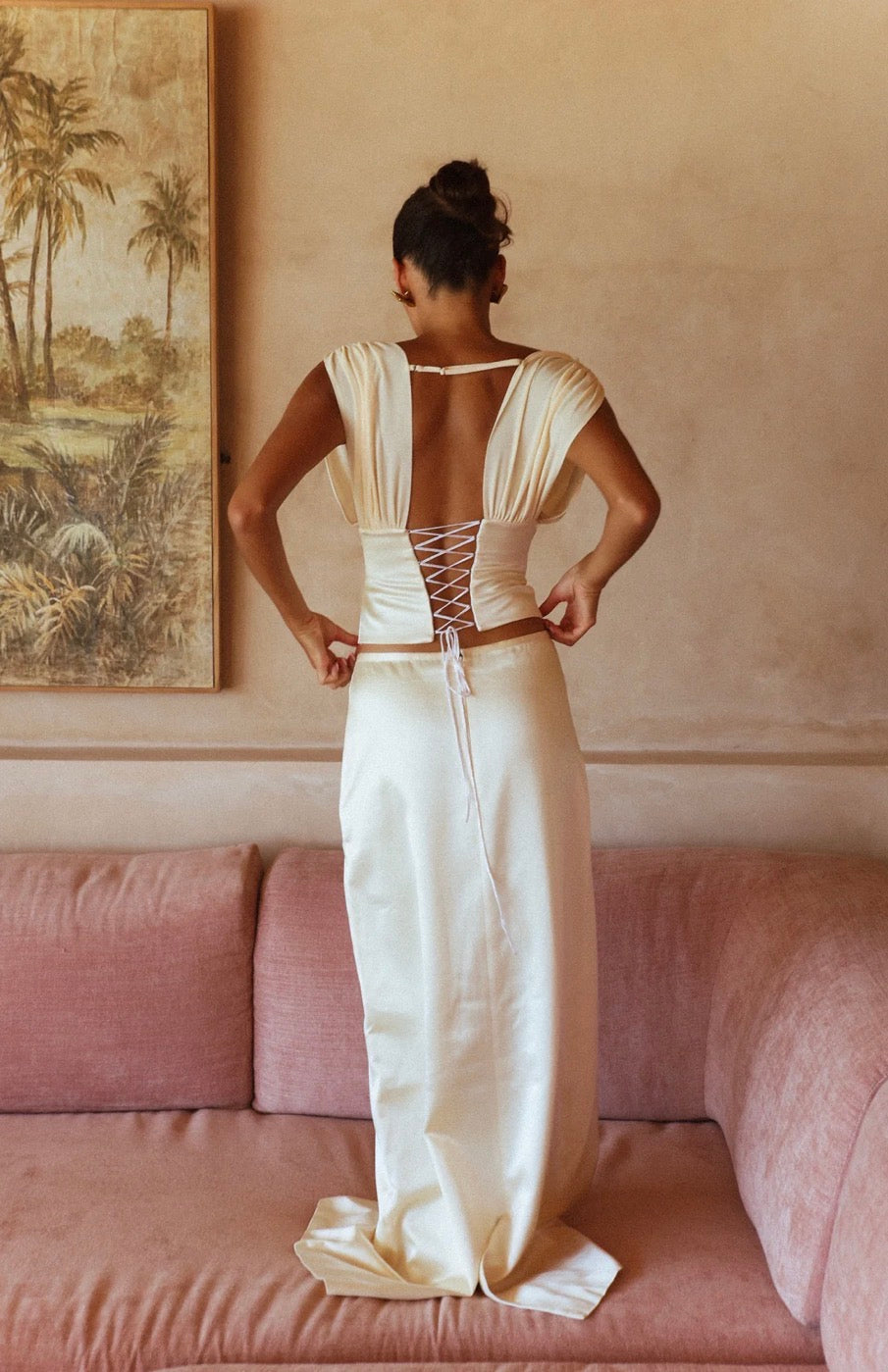 A woman stands with her back to the camera wearing a cream satin gown with a deep open back and lace-up detailing. She is posed in front of a dusty pink sofa with a framed landscape painting on the beige wall behind her.