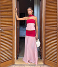 A woman stands in an open doorway wearing a two-piece pink and red dress with a halter top and a long flowing skirt. She is holding a white textured handbag and posing in front of wooden shutters. - @ella_squire