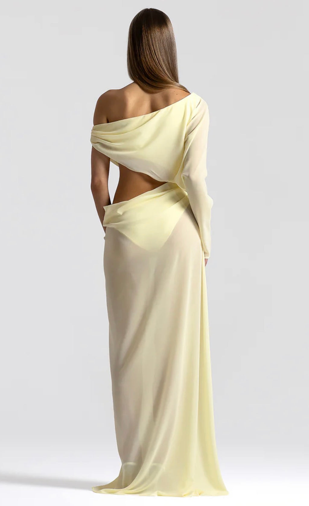 A woman shown from the back wearing a flowing one-shoulder maxi dress in pale pastel yellow, photographed against a light gray studio background.