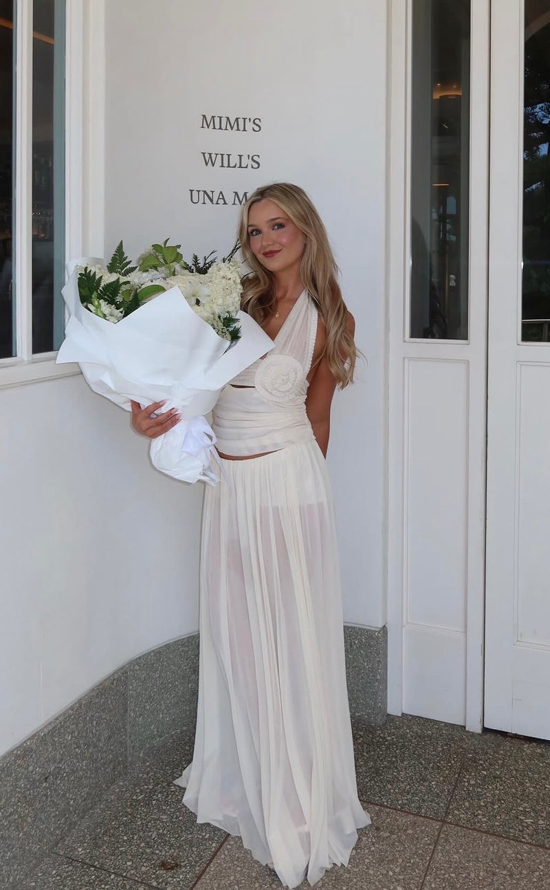 A woman stands outdoors in front of a white building door holding a large bouquet of white flowers, wearing a sleeveless white maxi dress with a draped waist and flowing skirt. - @chesleabramichh