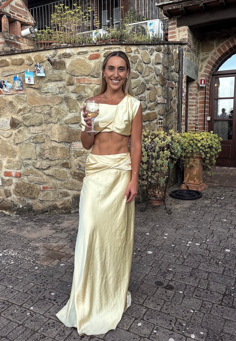 A woman stands outdoors in front of a rustic stone wall, wearing a pale yellow satin two-piece outfit with a cropped draped top and a long draped skirt. She is holding a glass of red wine. - @kailiephillpot