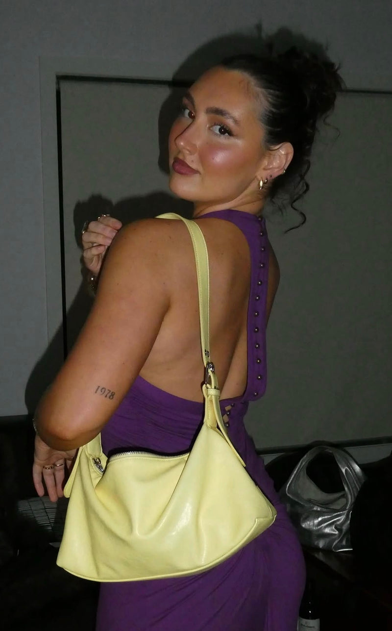 A woman wearing a purple dress and carrying a large pale yellow shoulder bag, photographed in a dim indoor setting. - @lilyrosetaylorr