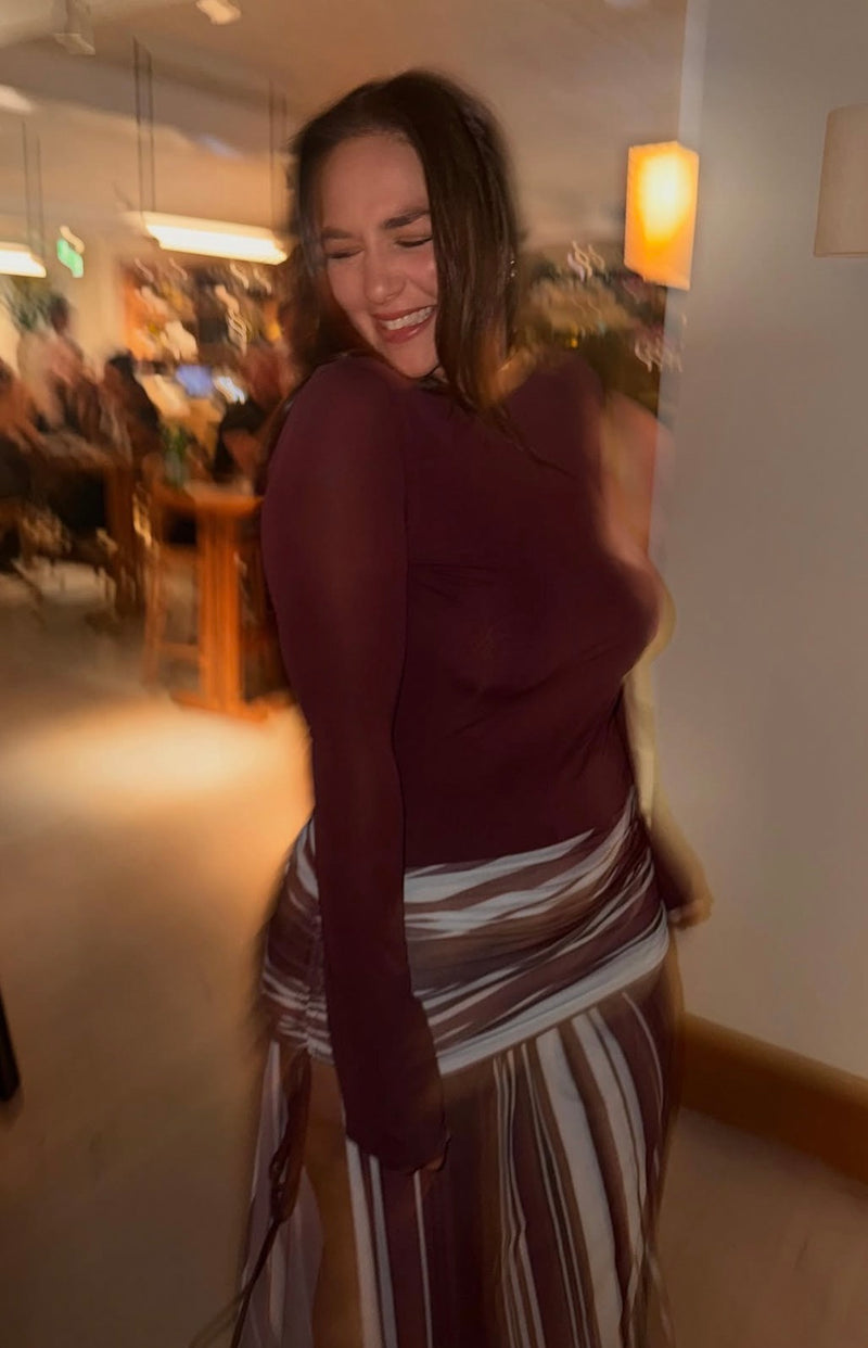 A woman wearing a sheer dark burgundy long-sleeve top and a brown, white, and beige striped skirt, smiling and posing in a warmly lit indoor setting. - @sarahfinlaay
