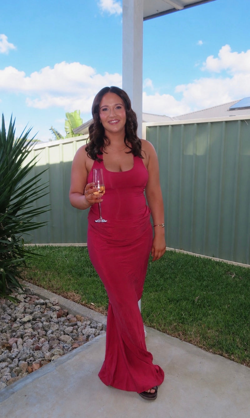 Girl standing in front of green fence with a wine glass in her hand wearing a red maxi dress smiling. - @chanaiduncombe