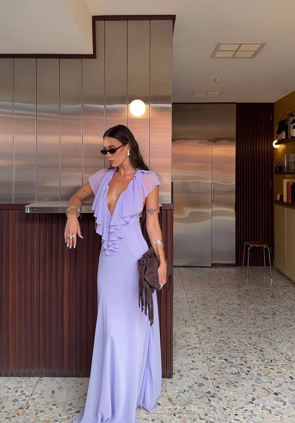A woman wearing a lavender purple ruffled maxi dress with a plunging neckline, standing in a modern interior with metallic paneling and terrazzo flooring. - @rahneebransby