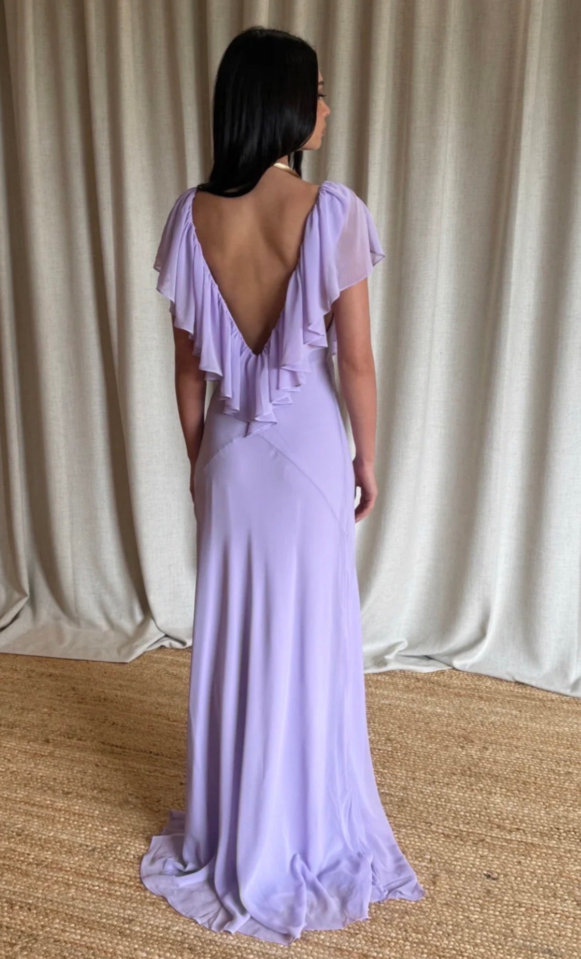 A woman wearing a lavender purple maxi dress with a ruffled open back and short flutter sleeves, shown from behind against a beige curtain backdrop.
