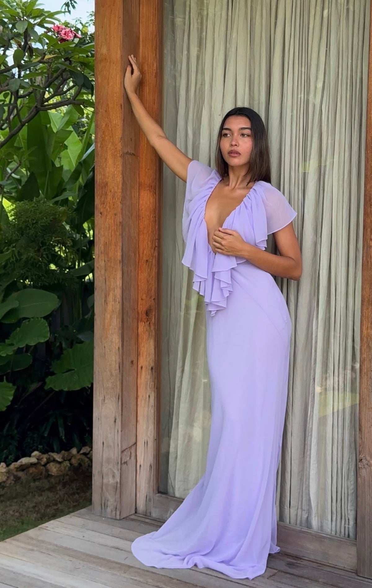A woman models a lavender purple maxi dress with a ruffled off-the-shoulder neckline and a deep V-neckline, standing on a wooden deck in front of a rustic wooden door with sheer curtains and green foliage in the background.