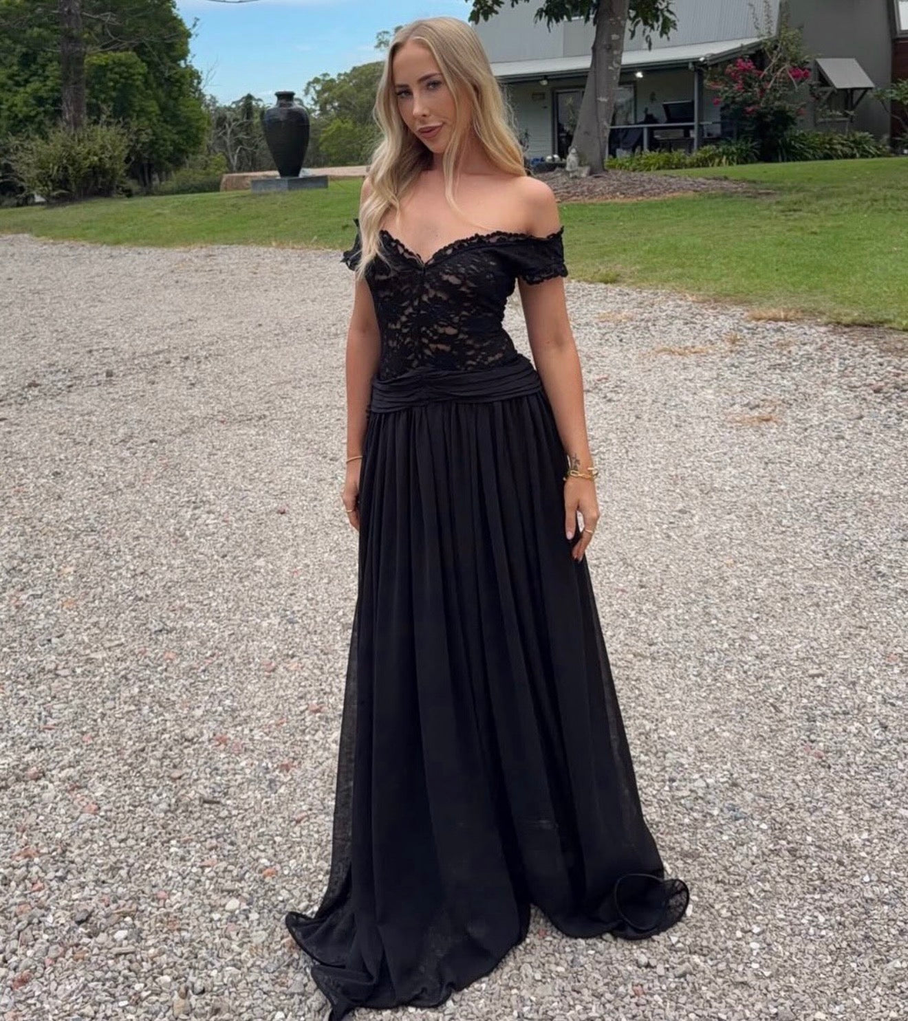 Woman in a black off-shoulder evening gown standing on a gravel path with greenery in the background. - @kaitlinknightt
