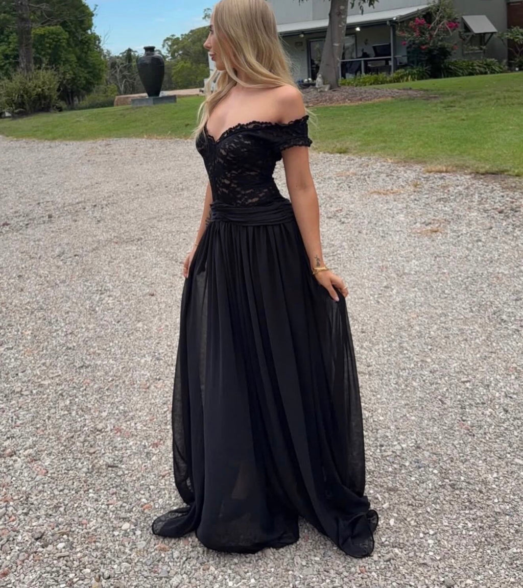 Woman wearing a black off-shoulder gown standing on a gravel path with grass and trees in the background.