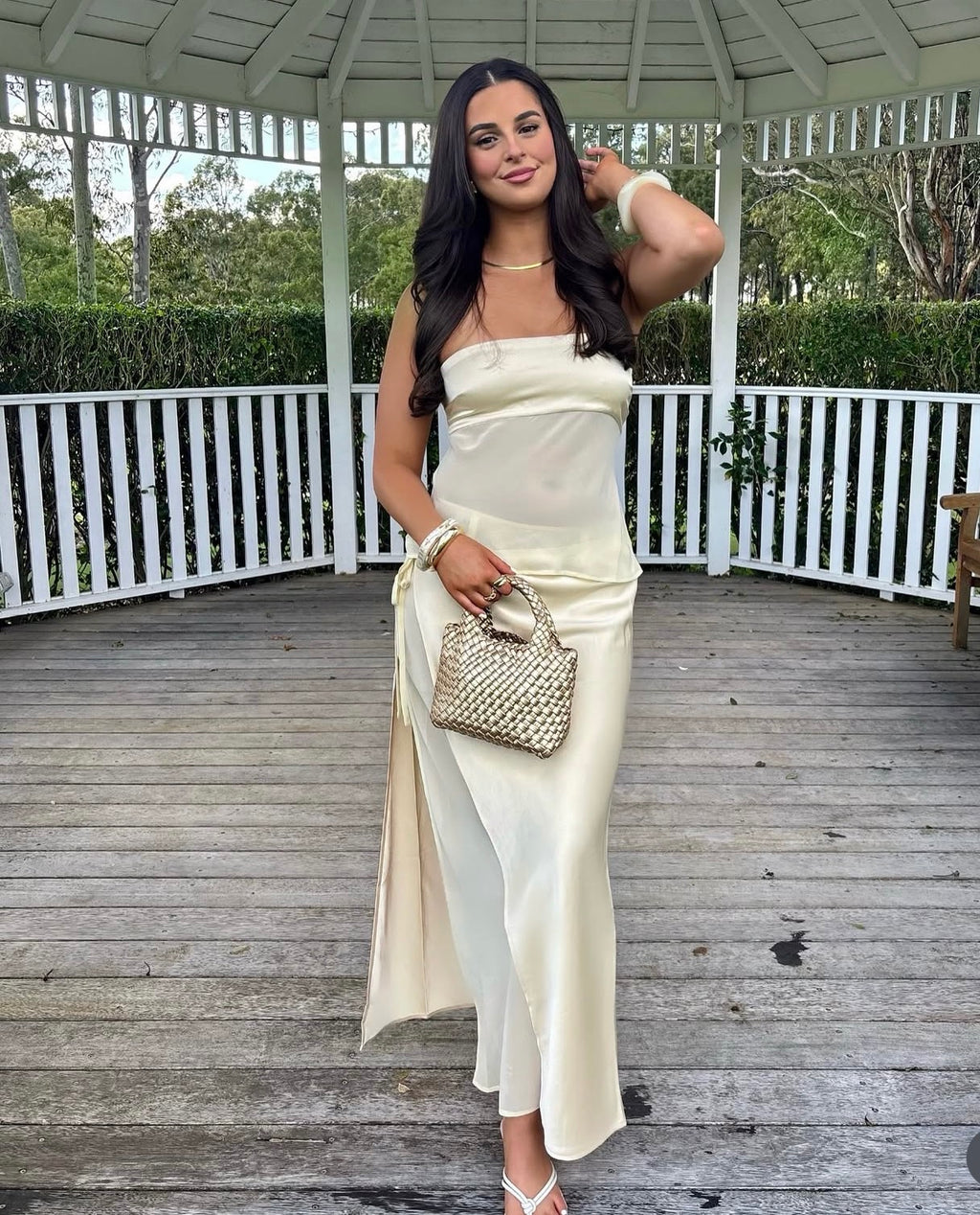 Woman in a white strapless dress holding a handbag on a wooden deck with a gazebo in the background.