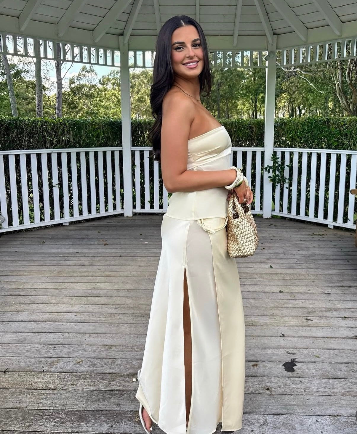 Woman in a light-colored outfit standing on a wooden deck with a white railing and greenery in the background. - @courtneyhoffman