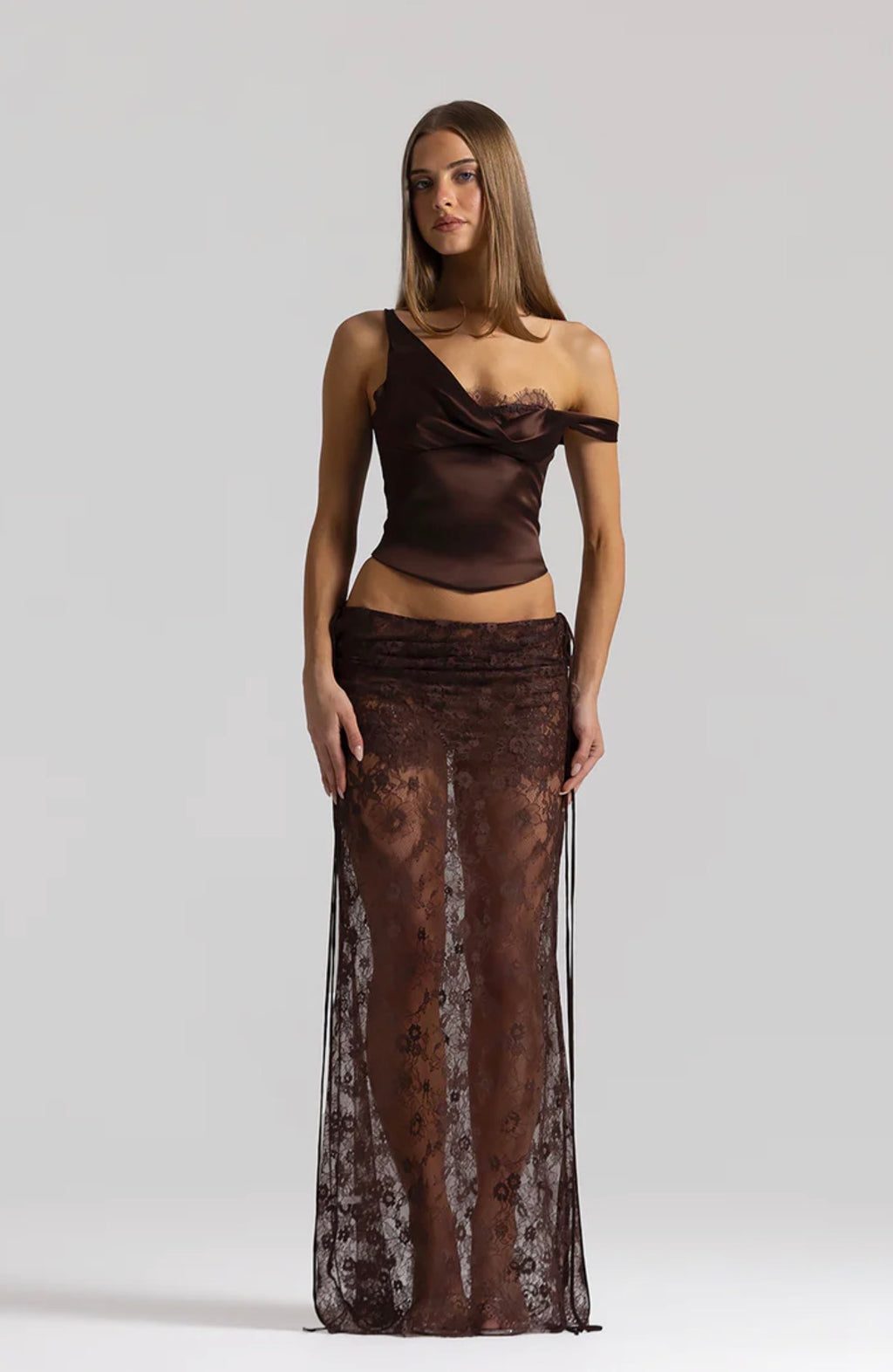 A model wearing a dark brown two-piece evening gown set, featuring a satin off-the-shoulder top and a matching sheer lace maxi skirt, against a light gray studio background.