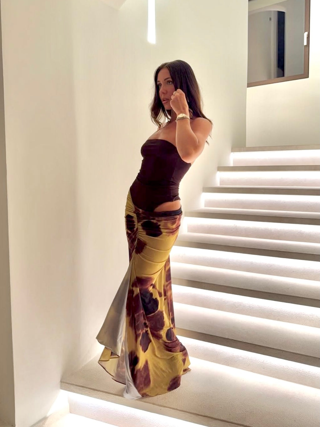 Girl standing in front of stairs with a white background wearing a brown dress with floral print skirt.