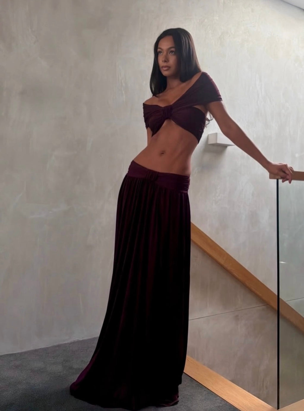 A woman modeling a deep burgundy two-piece outfit consisting of a short-sleeve wrap-style crop top and a matching pleated maxi skirt, standing in an indoor setting with a textured wall and glass railing.