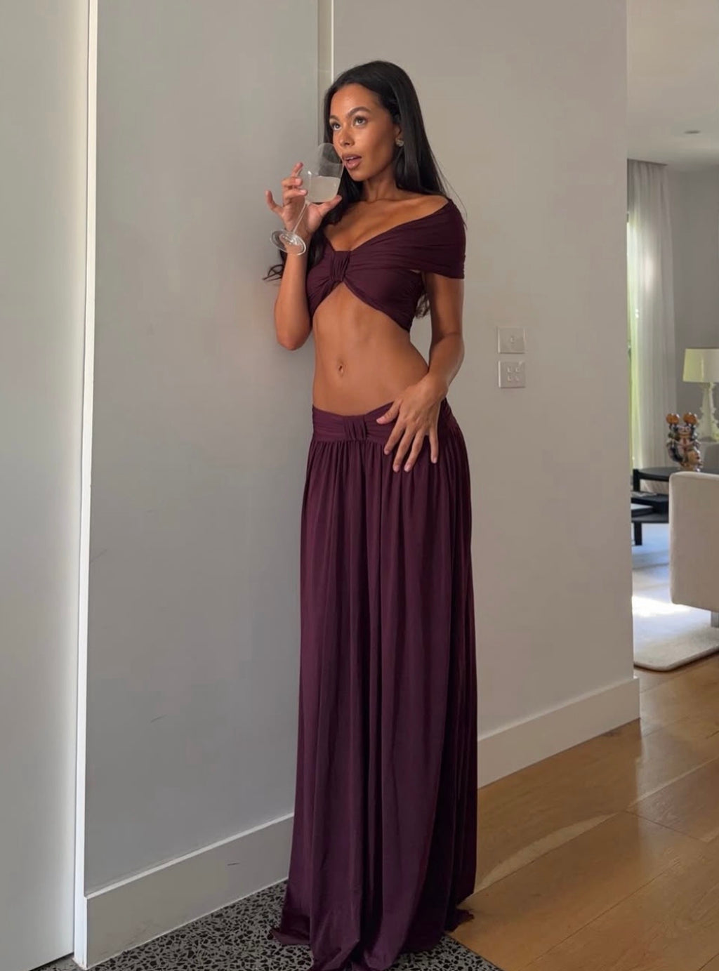 A woman wearing a two-piece deep plum purple dress set, consisting of a wrap-style off-the-shoulder crop top and a matching high-waisted maxi skirt, standing in a home hallway.