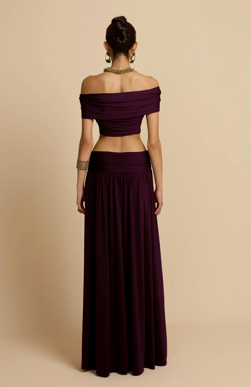 A woman shown from the back wearing a deep plum off-the-shoulder two-piece outfit with a cropped top and a long pleated skirt, against a solid beige studio background.