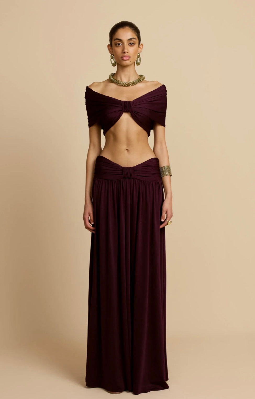 A model wearing a deep burgundy two-piece dress with a cropped off-the-shoulder top featuring a front twist detail and a matching high-waisted pleated skirt, set against a solid beige background.