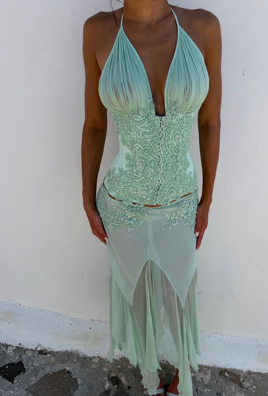 Women wearing a beaded halter green top paired with a sheer skirt standing pavement in front of a white wall.