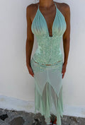 Women wearing a beaded halter green top paired with a sheer skirt standing pavement in front of a white wall.
