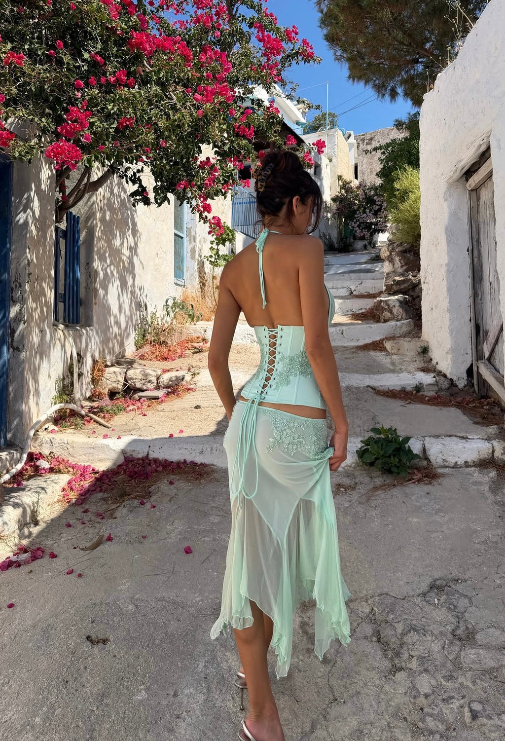 A woman wearing a mint green halter-neck maxi dress with lace-up back detailing and a sheer overlay skirt, walking down a sunlit stone alleyway with white buildings and pink flowers.