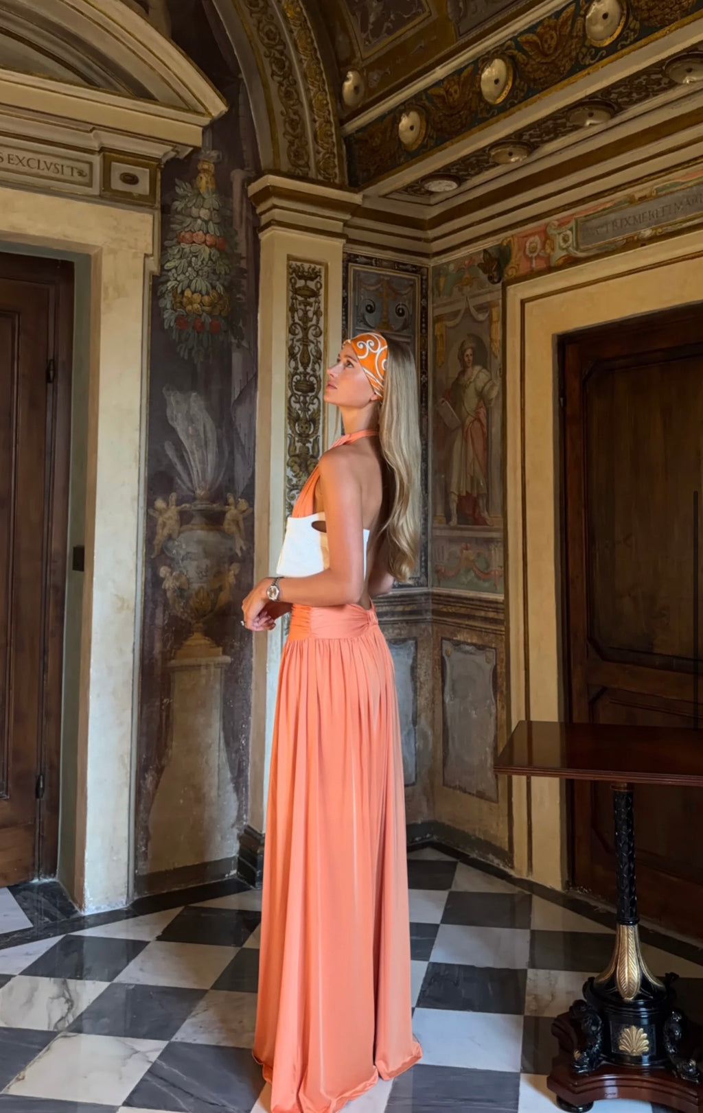 A woman stands in profile in an ornate interior hallway with painted walls, a checkered floor, and a large peach maxi dress. She is holding a white clutch and wearing a matching peach headband.