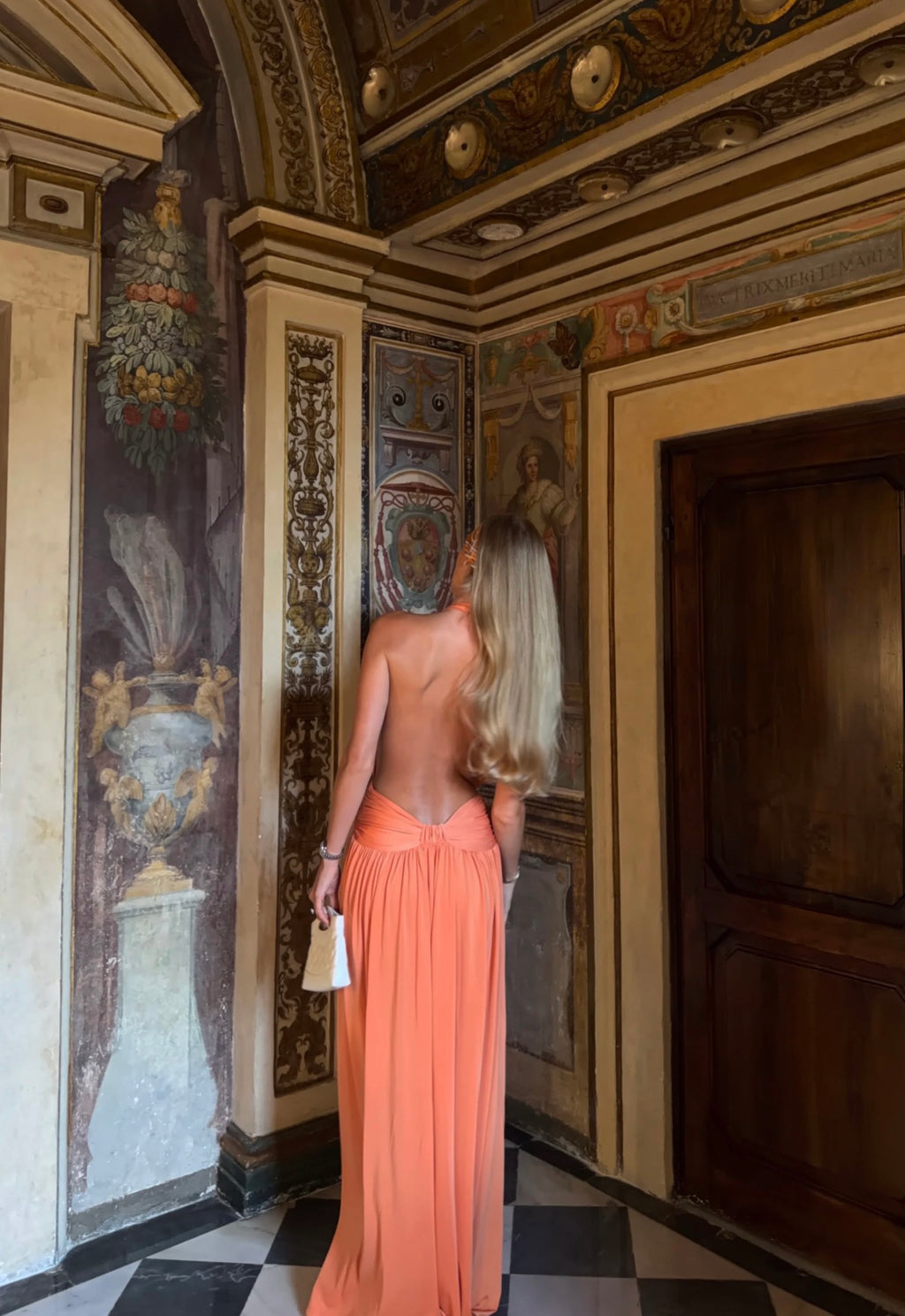 A woman with long blonde hair stands in a grand, ornate interior hallway wearing a flowing peach-colored maxi dress with a low back. She is seen from behind, holding a small white handbag.