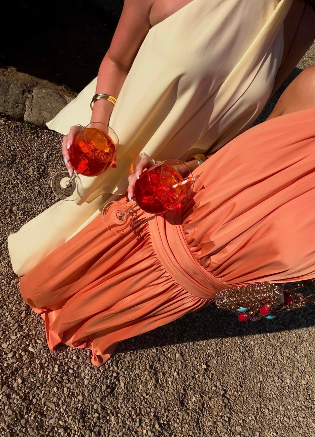 A close-up lifestyle shot of a person wearing a cream one-shoulder dress and a peach ruched skirt, holding two glasses of red cocktail on a gravel surface.