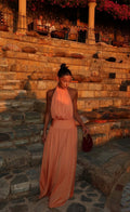 A woman wearing a peach halter-neck maxi dress walks down stone steps at sunset, holding a small dark red handbag.
