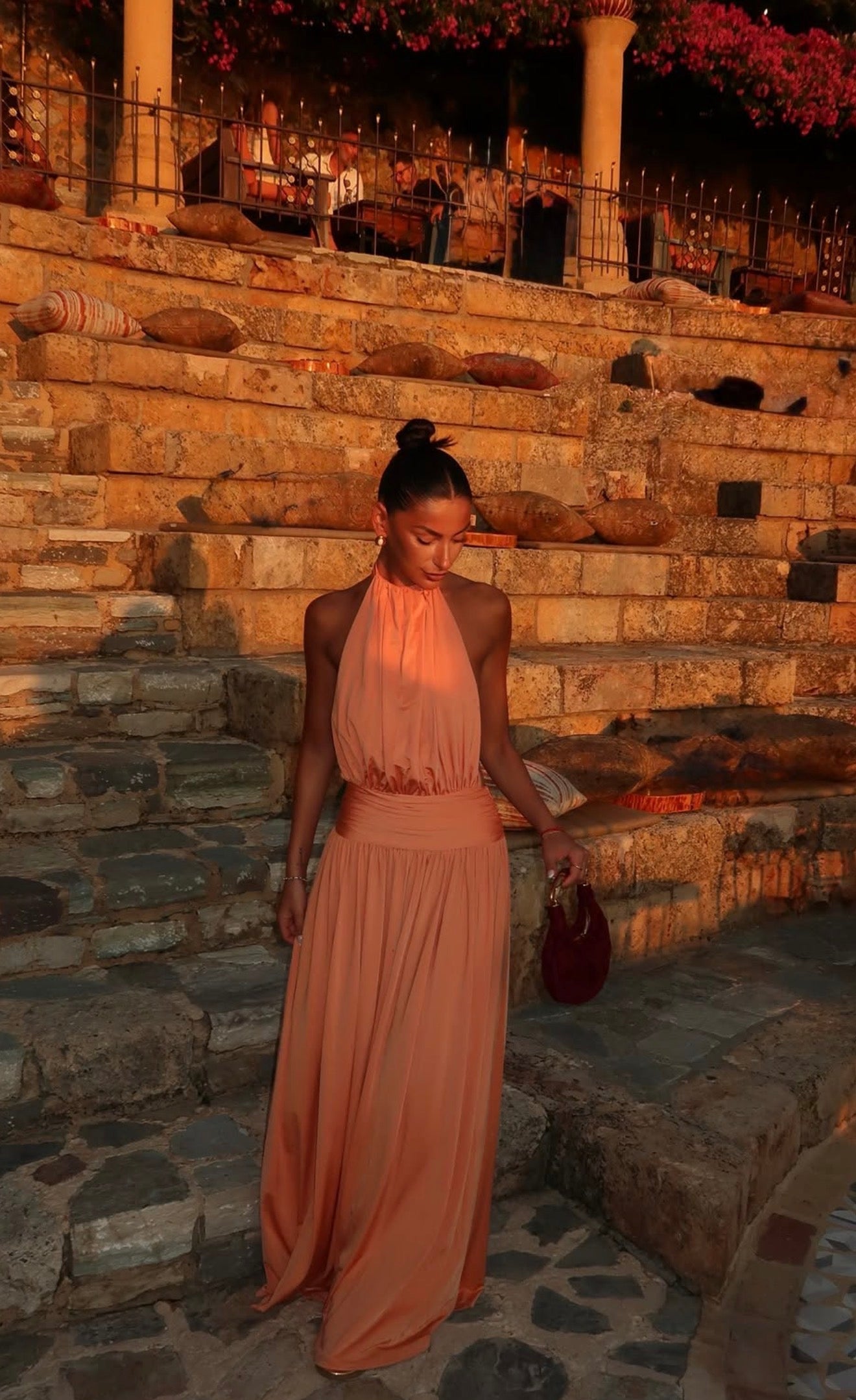 A woman wearing a peach halter-neck maxi dress walks down stone steps at sunset, holding a small dark red handbag.