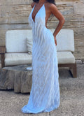 A woman wearing a light blue feather maxi dress with a deep V-neckline and back, standing in front of a rustic wooden wall and a beige outdoor sofa.