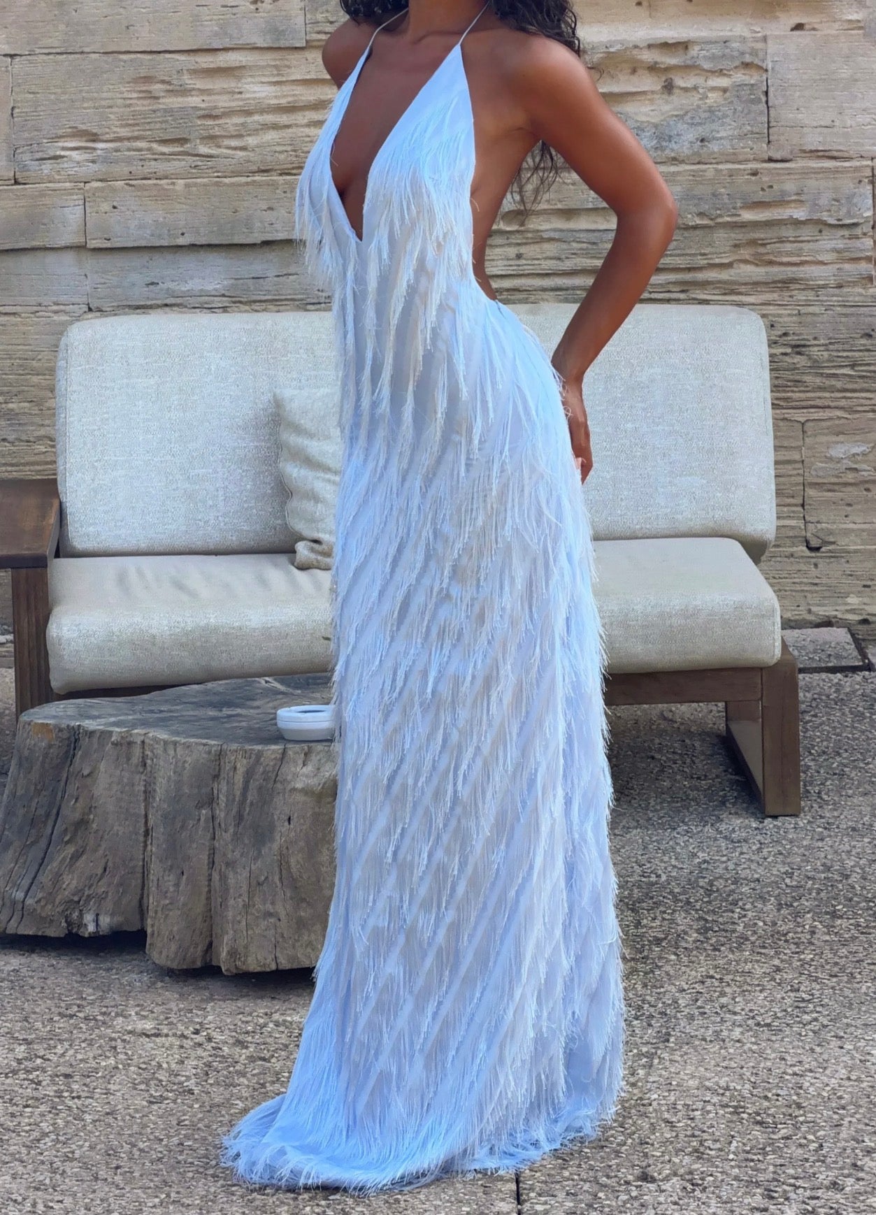A woman wearing a light blue feather maxi dress with a deep V-neckline and back, standing in front of a rustic wooden wall and a beige outdoor sofa.