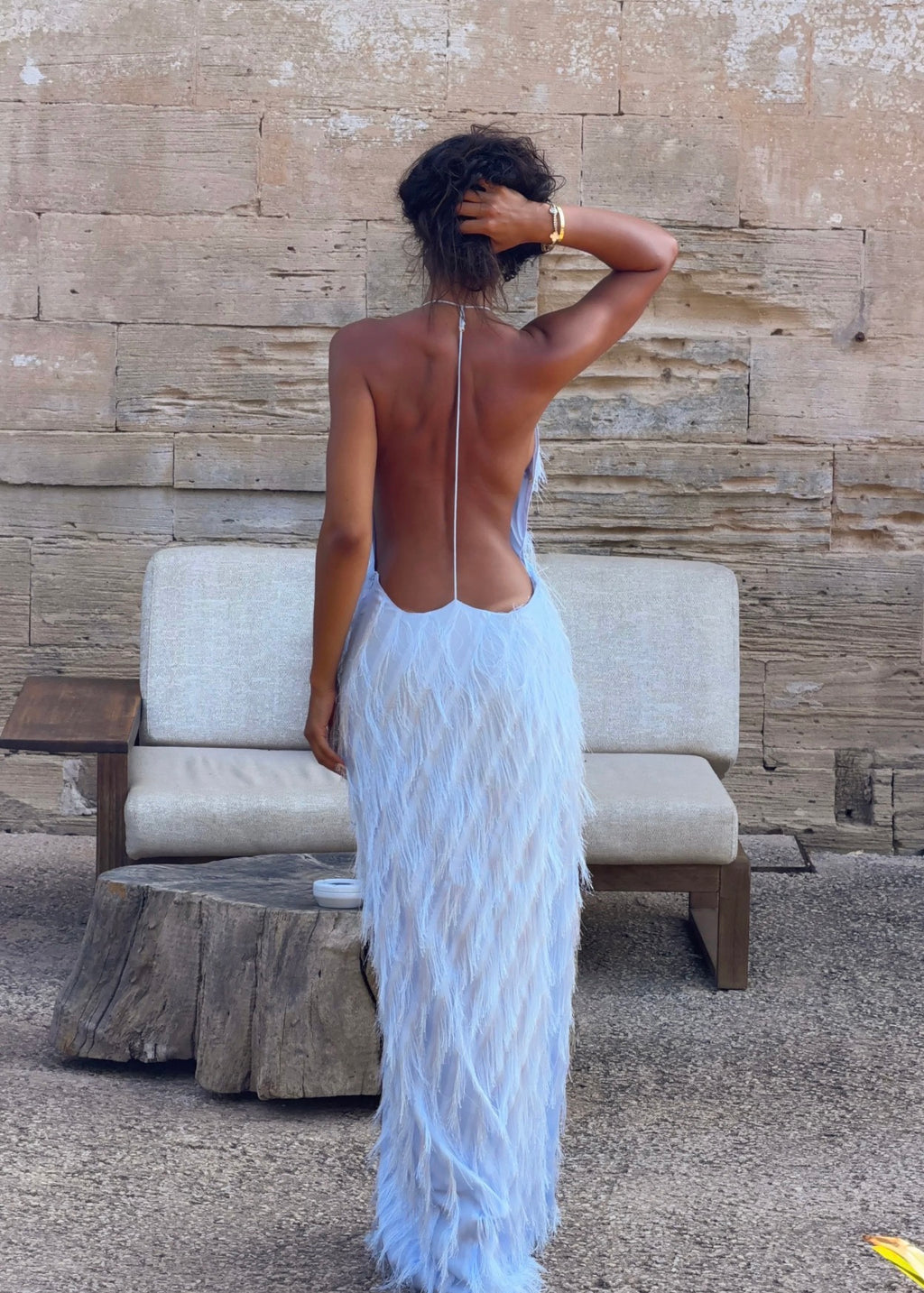 A woman stands with her back to the camera wearing a light blue feathered maxi dress with an open back, standing in front of a rustic stone wall and a beige lounge chair.