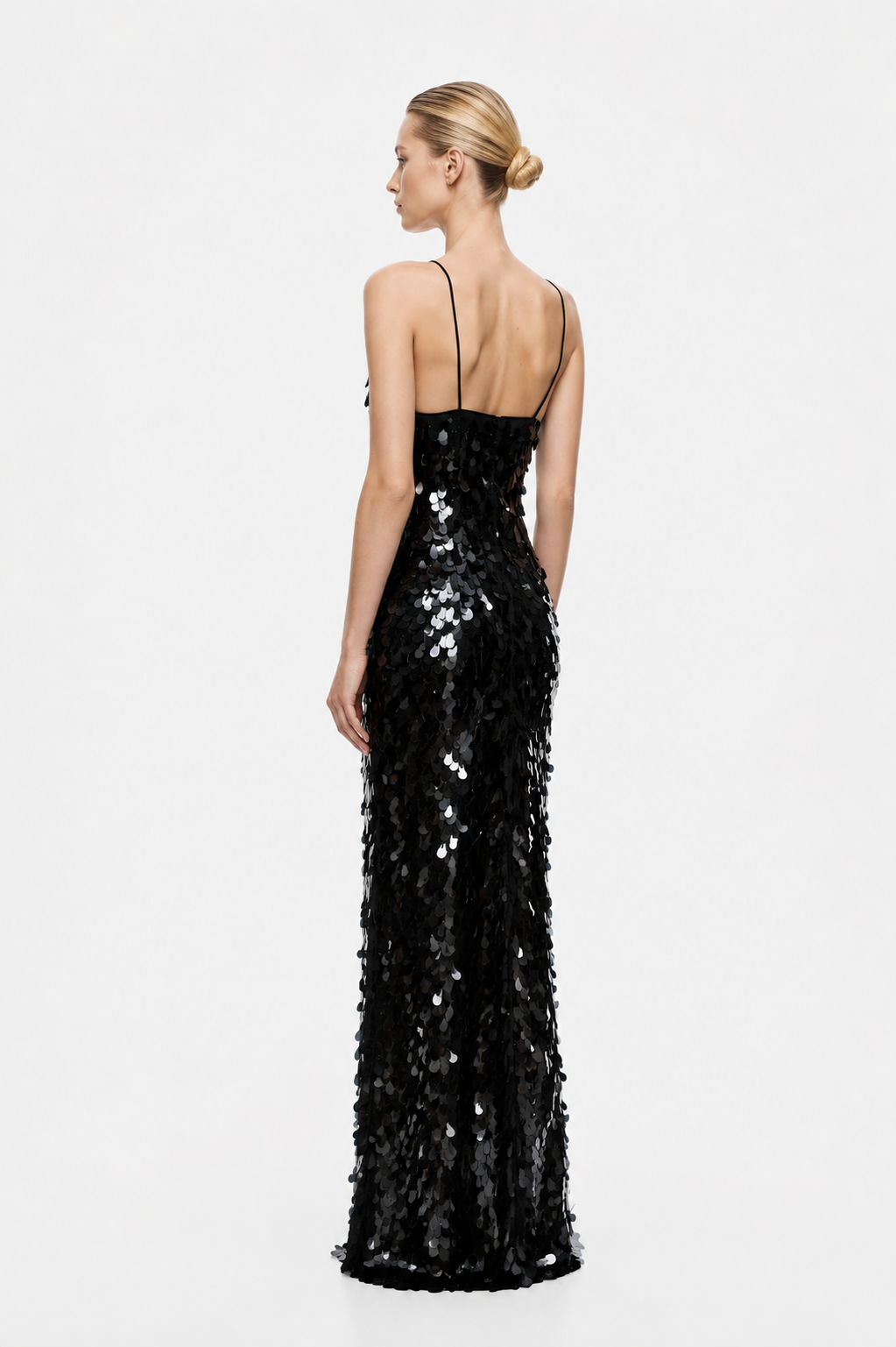 A woman modeling a floor-length black sequined gown from the back against a plain light gray studio background.
