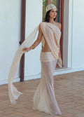 A woman models a draped, one-shoulder cream chiffon dress with a high open back and a matching sheer wrap skirt, styled with a beaded headband in an outdoor patio setting.