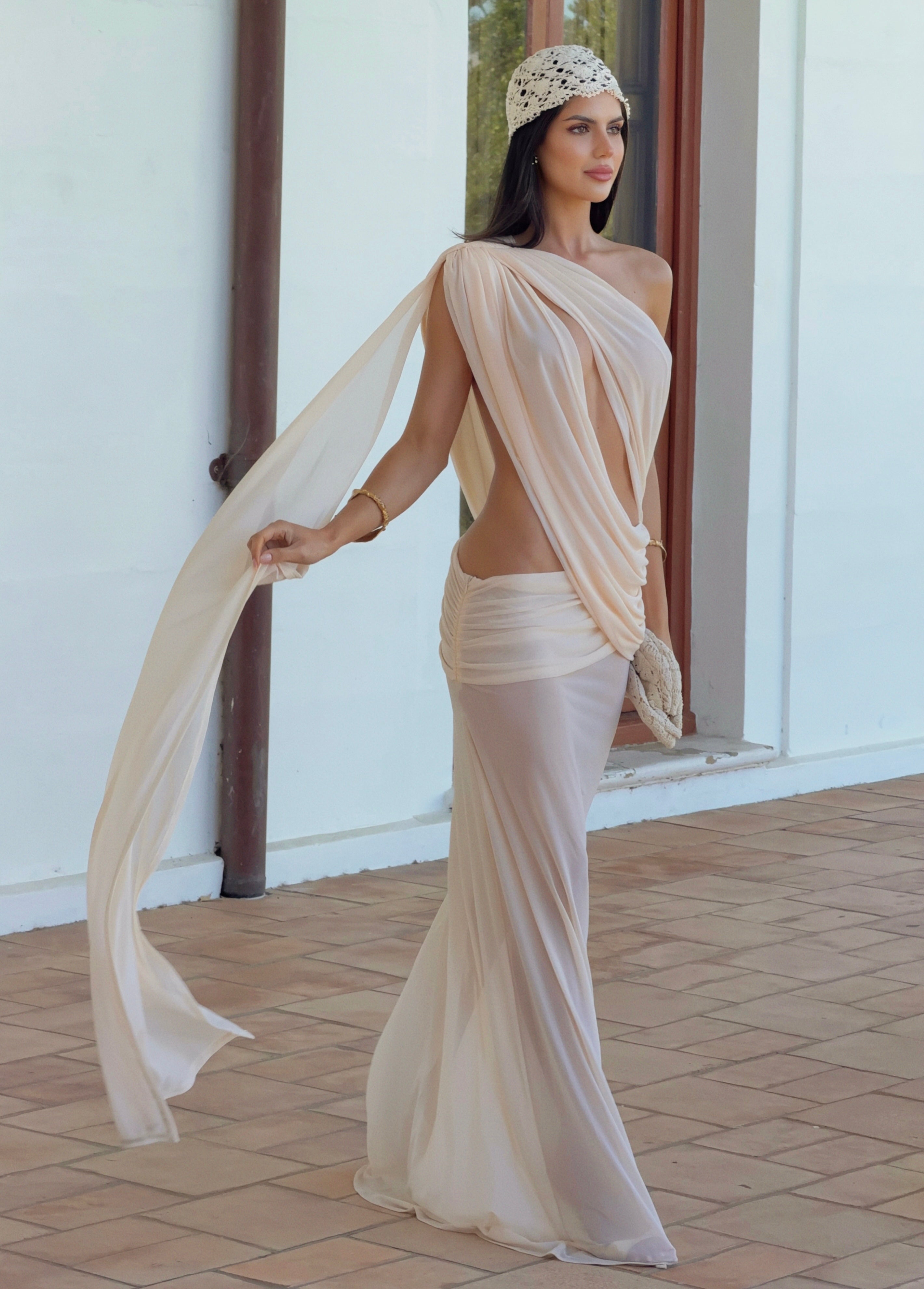 A woman models a draped, one-shoulder cream chiffon dress with a high open back and a matching sheer wrap skirt, styled with a beaded headband in an outdoor patio setting.