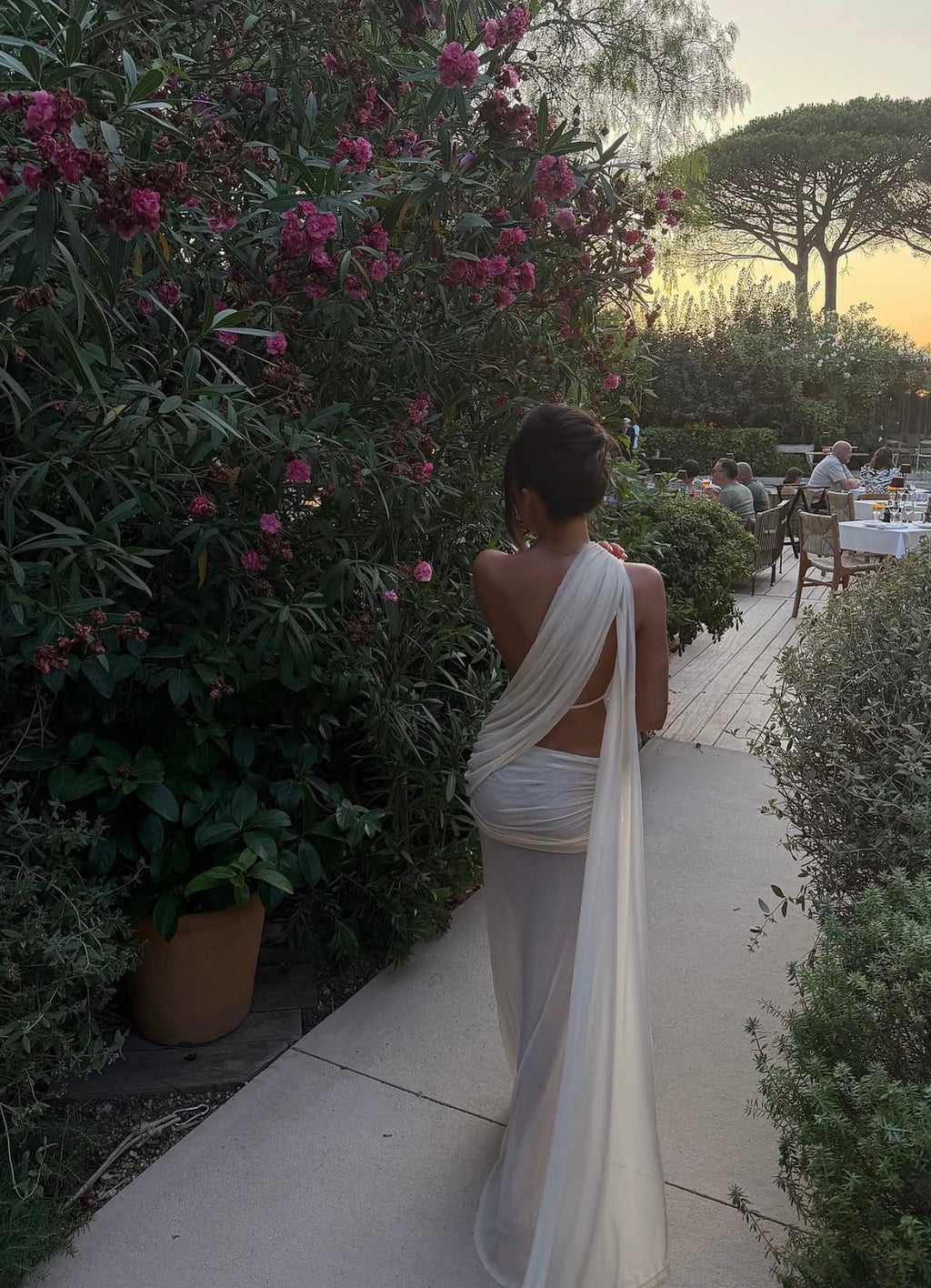 A woman viewed from behind wearing a flowing white draped dress or sari, standing on a garden path beside dense greenery and pink flowers at dusk.
