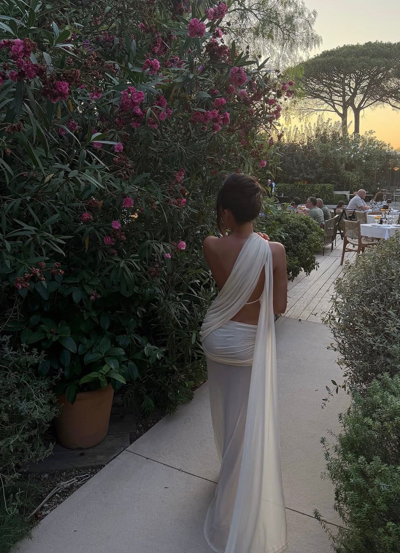 A woman viewed from behind wearing a flowing white draped dress or sari, standing on a garden path beside dense greenery and pink flowers at dusk.