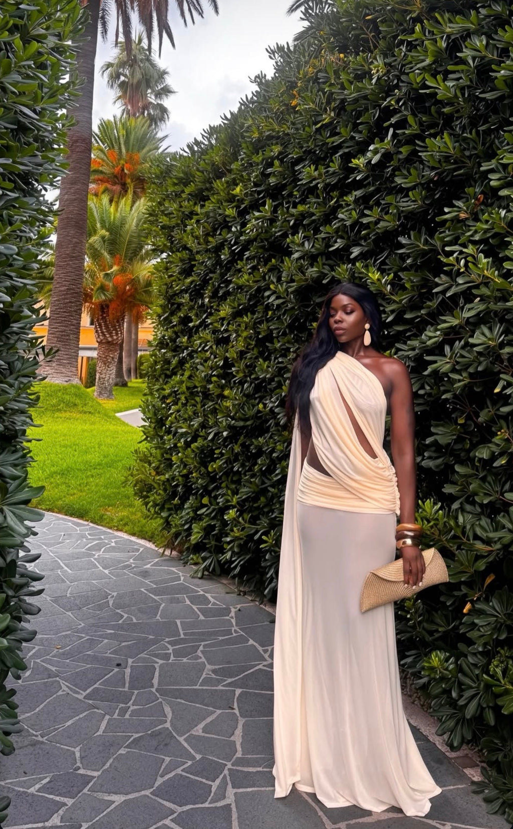 A woman stands outdoors on a stone path beside a tall green hedge, wearing a flowing one-shoulder cream maxi dress with a draped neckline and side cutout. She holds a small beige clutch and wears statement earrings and bracelets.