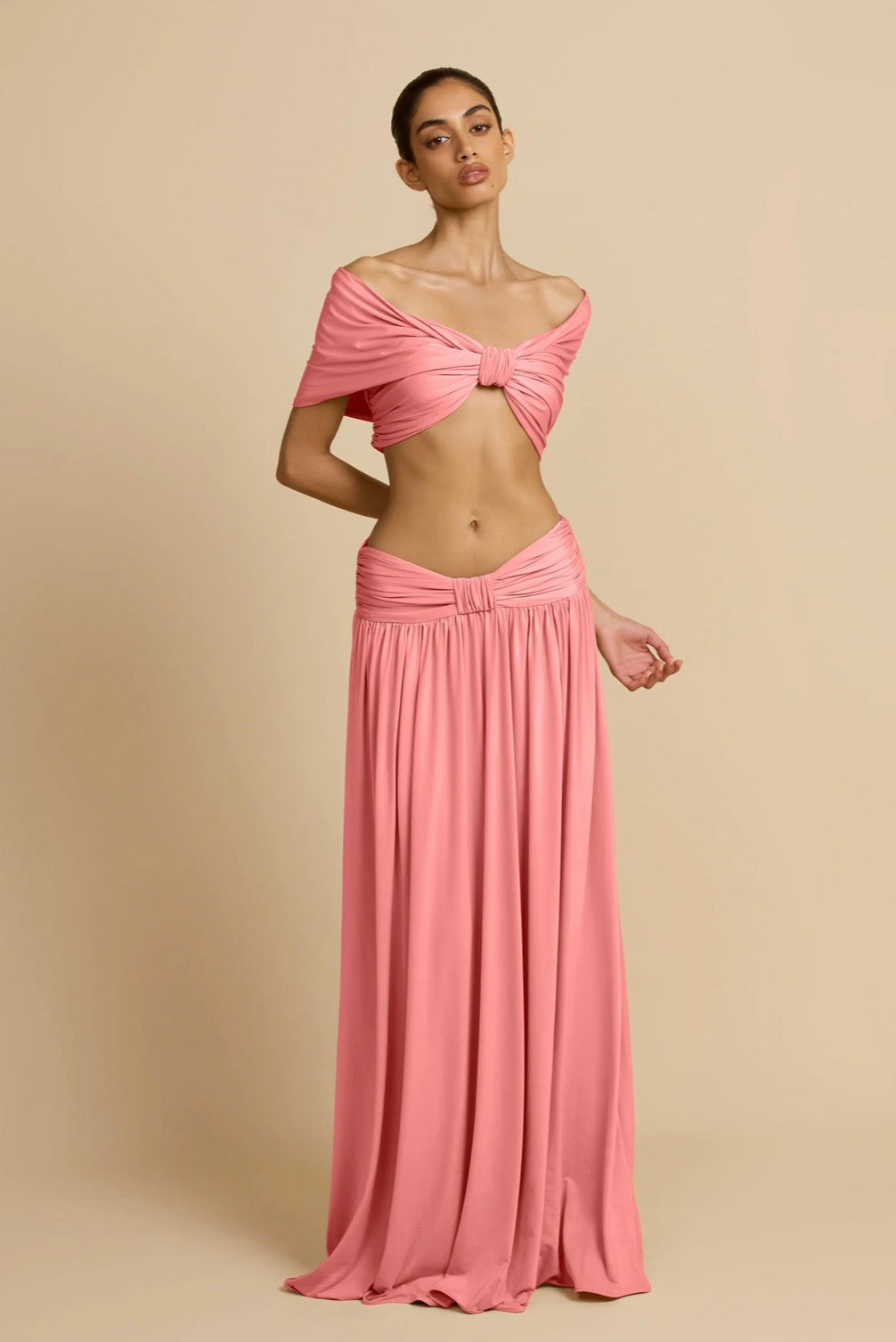 A model wearing a two-piece pink set consisting of a cropped off-the-shoulder top with a front twist detail and a matching high-waisted pleated maxi skirt, set against a solid beige studio background.