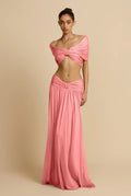 A model wearing a two-piece pink formal gown set consisting of an off-the-shoulder cropped top with a front twist detail and a matching floor-length skirt, set against a solid beige studio background.