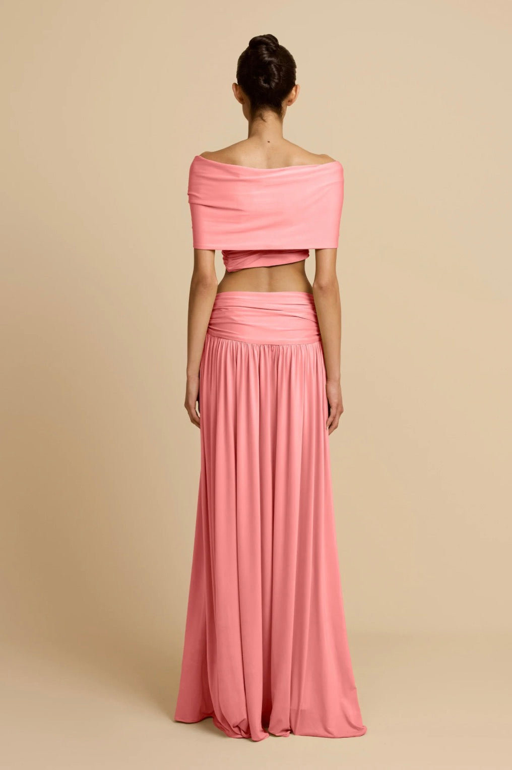 A model shown from the back wearing a two-piece pink outfit consisting of an off-the-shoulder crop top and a matching pleated maxi skirt, set against a solid beige background.