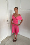 A woman modeling a bright pink two-piece set consisting of an off-the-shoulder wrap-style crop top and a matching flared mini skirt, paired with black flip-flop sandals and a gold necklace, standing in a white-walled room.