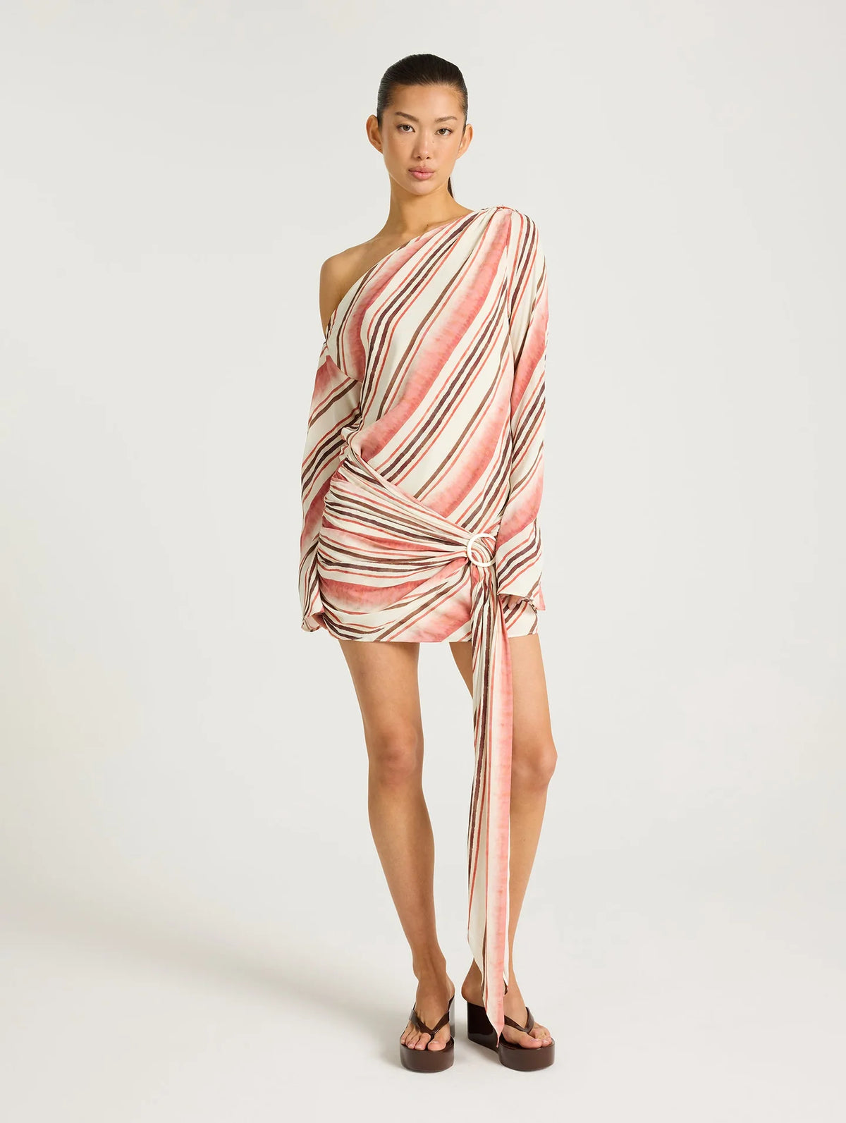 Model wearing a pink, cream and brown striped one-shoulder mini dress with draped detailing, long sleeve and tie feature at the hip.