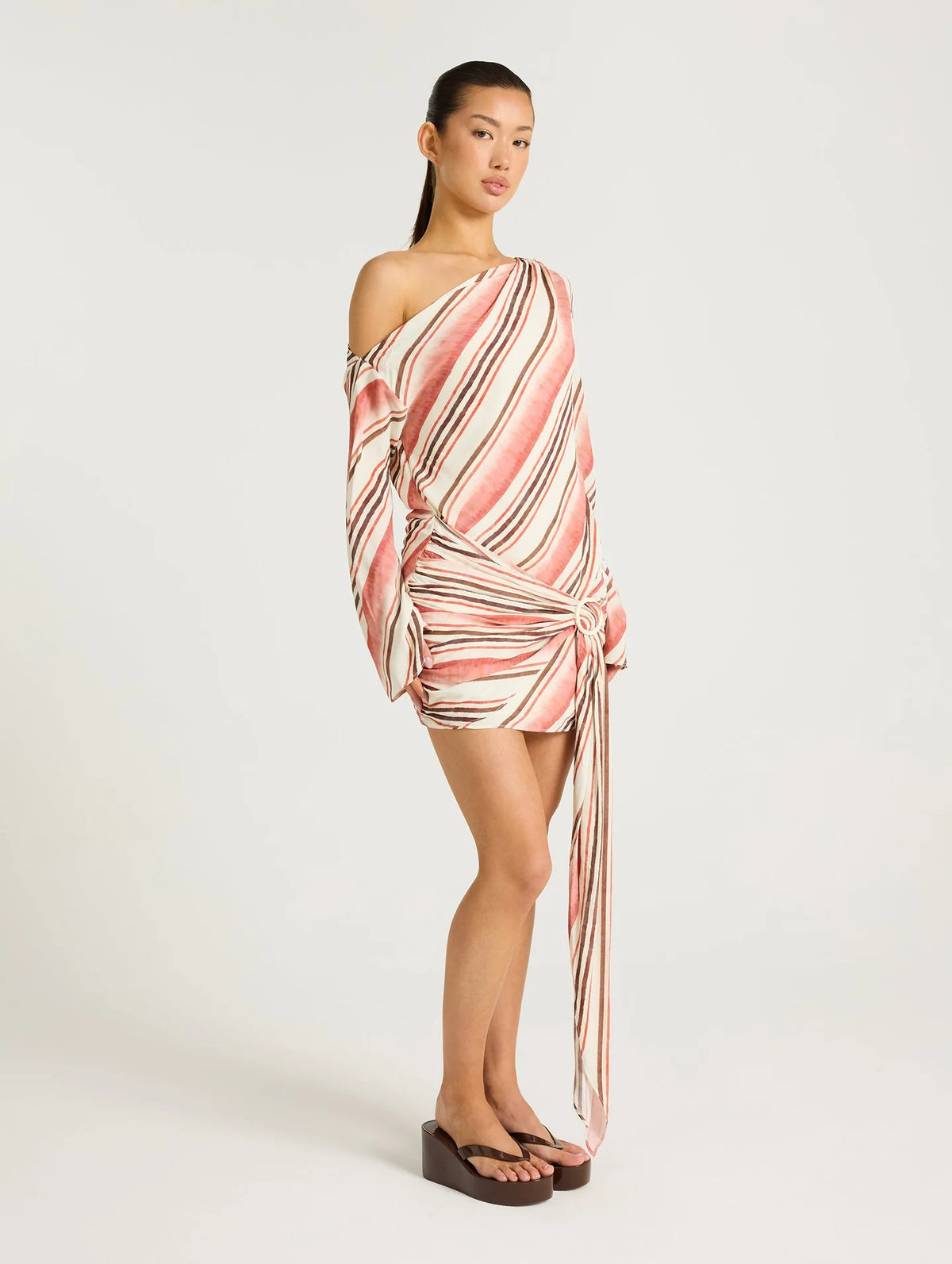 A model wearing a one-shoulder wrap mini dress with diagonal pink, beige, and white stripes, styled with dark brown wedge sandals against a plain light gray studio background.